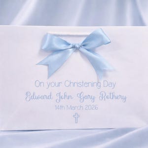 Op de afbeelding: Een witte cadeauzak met een lichtblauwe satijnen strik aan de bovenkant. De tekst luidt On your Christening Day, Edward John Gary Rothery, 14th March 2026, met een klein lichtblauw kruissymbool onder de datum gedrukt in een eenvoudig lettertype.