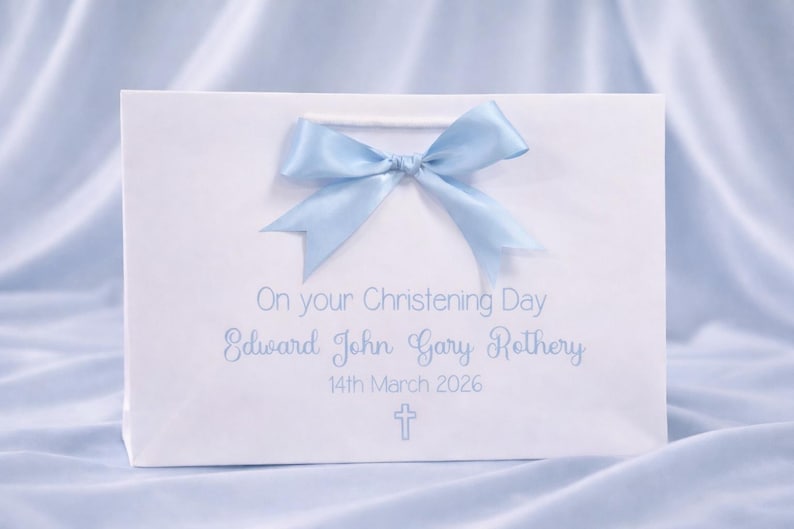 Op de afbeelding: Een witte cadeauzak met een lichtblauwe satijnen strik aan de bovenkant. De tekst luidt On your Christening Day, Edward John Gary Rothery, 14th March 2026, met een klein lichtblauw kruissymbool onder de datum gedrukt in een eenvoudig lettertype.