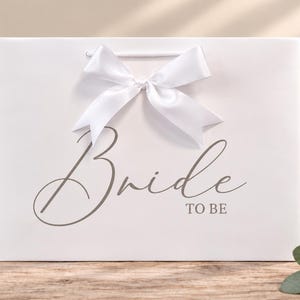 May include: A white rectangular gift bag featuring a large white satin ribbon bow at the top center. The front of the bag displays the text "Bride TO BE" in a dark gray script and sans-serif font combination centered on the surface.