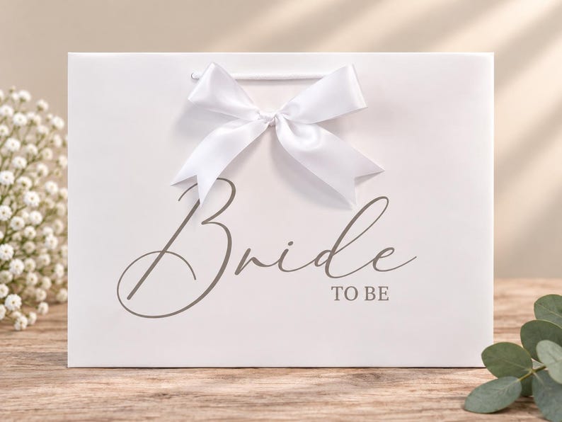 May include: A white rectangular gift bag featuring a large white satin ribbon bow at the top center. The front of the bag displays the text "Bride TO BE" in a dark gray script and sans-serif font combination centered on the surface.