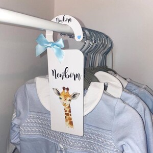 May include: A white hanger with a blue ribbon and a white card that says "newborn" with a giraffe illustration. The card is hanging on a white rod with several other hangers.