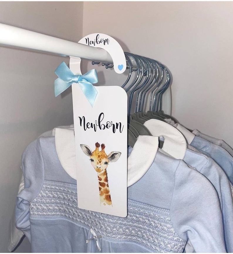 May include: A white hanger with a blue ribbon and a white card that says "newborn" with a giraffe illustration. The card is hanging on a white rod with several other hangers.