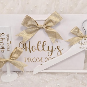 May include: A white gift bag, a white champagne flute, and a white clothes hanger, each featuring gold text that reads Holly's PROM 2026. The items are decorated with shimmering gold ribbon bows, creating a coordinated set for a special event.