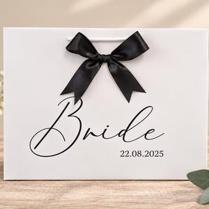 May include: A white rectangular gift bag featuring a black satin ribbon bow at the top center. The word Bride is printed in a black cursive script font, with the date 22.08.2025 printed in a smaller black serif font below it.