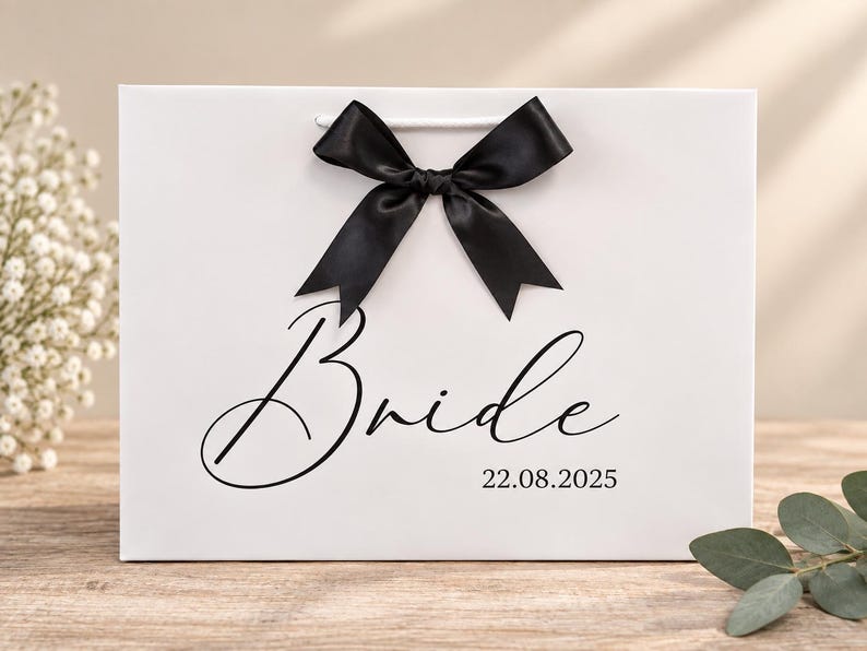 May include: A white rectangular gift bag featuring a black satin ribbon bow at the top center. The word Bride is printed in a black cursive script font, with the date 22.08.2025 printed in a smaller black serif font below it.