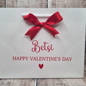 May include: White gift bag with a red bow and the text "Betsi Happy Valentine's Day" in red lettering with a small red heart.