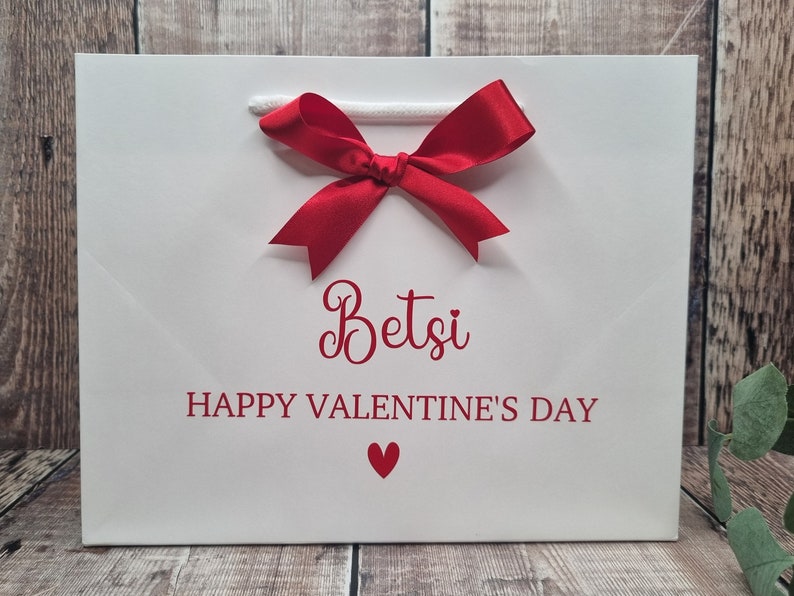 May include: White gift bag with a red bow and the text "Betsi Happy Valentine's Day" in red lettering with a small red heart.