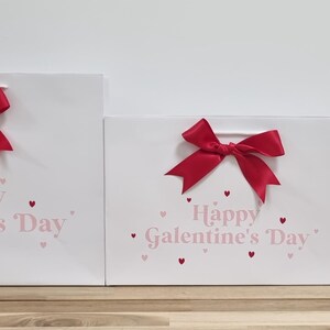 May include: Three white gift bags with red bows and pink hearts. The bags have the text "Happy Galentine's Day" printed on them.