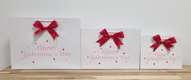 May include: Three white gift bags with red bows and pink hearts. The bags have the text "Happy Galentine's Day" printed on them.