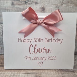 Puede incluir: Bolsa de regalo blanca con un lazo de sat&eacute;n rosa. La bolsa tiene el texto "Happy 50th Birthday Claire 17th January 2025" y un s&iacute;mbolo de coraz&oacute;n rosa. La bolsa tiene asas blancas.