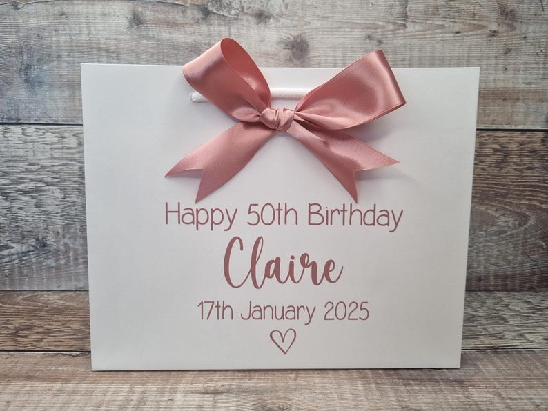 Puede incluir: Bolsa de regalo blanca con un lazo de sat&eacute;n rosa. La bolsa tiene el texto "Happy 50th Birthday Claire 17th January 2025" y un s&iacute;mbolo de coraz&oacute;n rosa. La bolsa tiene asas blancas.
