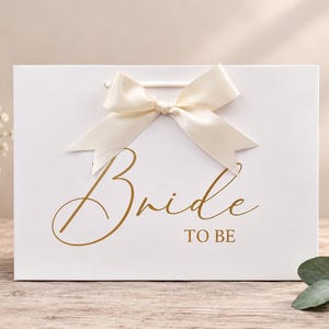 May include: A white rectangular gift bag featuring the gold text Bride TO BE in a script and block font combination. The bag is adorned with a cream-colored satin ribbon tied into a bow at the top center, positioned above the printed text.