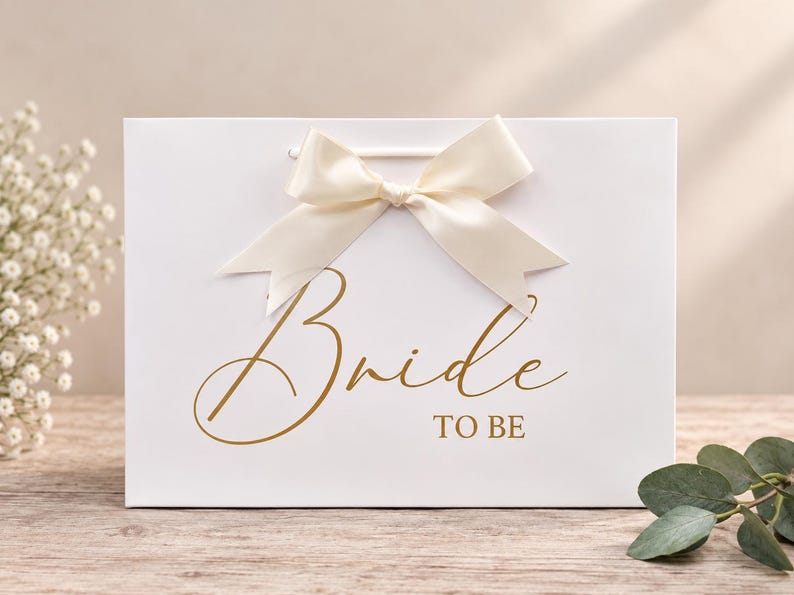 May include: A white rectangular gift bag featuring the gold text Bride TO BE in a script and block font combination. The bag is adorned with a cream-colored satin ribbon tied into a bow at the top center, positioned above the printed text.