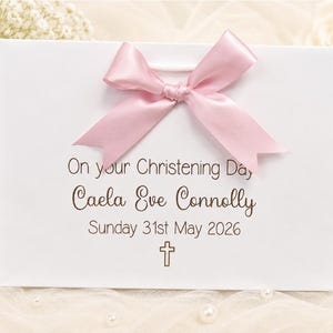 Op de afbeelding: Een witte rechthoekige cadeauzak met een lichtroze satijnen strik in het midden bovenaan. De tekst luidt: On your Christening Day, Caela Eve Connolly, Sunday 31st May 2026, met een klein kruissymbool in bruine inkt onder de datum.