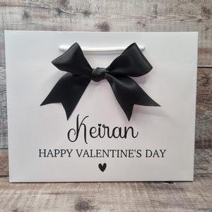 May include: White gift bag with a black bow and the text "Keiran Happy Valentine's Day" with a small heart.