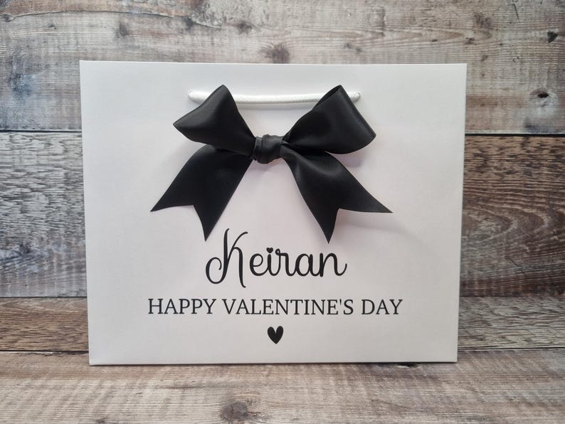May include: White gift bag with a black bow and the text "Keiran Happy Valentine's Day" with a small heart.