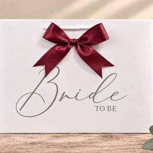 May include: A white rectangular gift bag featuring a dark burgundy satin ribbon bow centered at the top. The front of the bag displays the text "Bride TO BE" in a grey script and block font combination, with white cord handles.