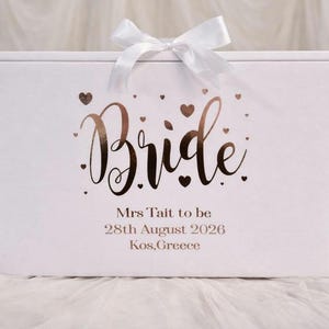 May include: A rectangular white gift box featuring a white satin ribbon bow on top. The front displays the word Bride in metallic rose gold script, surrounded by small hearts. Below, the text reads Mrs Tait to be, 28th August 2026, Kos, Greece.