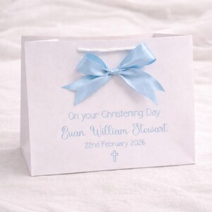 Op de afbeelding: Een witte cadeauzak met een lichtblauwe satijnen strik aan de bovenkant. De zak is bedrukt met de tekst On your Christening Day, de naam Euan William Stewart, de datum 22nd February 2026 en een klein lichtblauw kruissymbool.