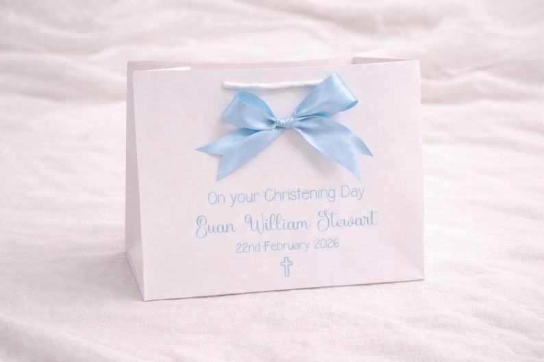 Op de afbeelding: Een witte cadeauzak met een lichtblauwe satijnen strik aan de bovenkant. De zak is bedrukt met de tekst On your Christening Day, de naam Euan William Stewart, de datum 22nd February 2026 en een klein lichtblauw kruissymbool.