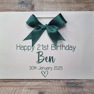 Puede incluir: Bolsa de regalo blanca con un lazo de sat&eacute;n verde oscuro. La bolsa tiene el texto "Happy 21st Birthday Ben" y la fecha "30 de enero de 2025" en verde oscuro, con un s&iacute;mbolo de coraz&oacute;n debajo.