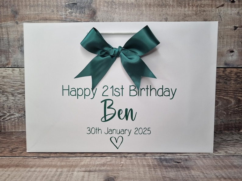 Puede incluir: Bolsa de regalo blanca con un lazo de sat&eacute;n verde oscuro. La bolsa tiene el texto "Happy 21st Birthday Ben" y la fecha "30 de enero de 2025" en verde oscuro, con un s&iacute;mbolo de coraz&oacute;n debajo.