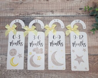 Puede incluir: Cuatro colgadores de hitos para bebés blancos con lazos amarillos y diferentes diseños de luna y estrellas. Los colgadores están etiquetados con los rangos de edad de 0 a 3 meses, de 3 a 6 meses, de 6 a 9 meses y de 9 a 12 meses.