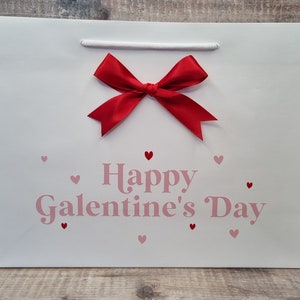 May include: White gift bag with a red bow and pink hearts. The bag says "Happy Galentine's Day".