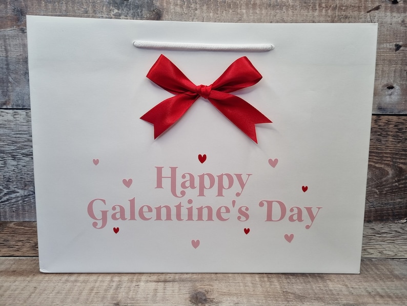 May include: White gift bag with a red bow and pink hearts. The bag says "Happy Galentine's Day".