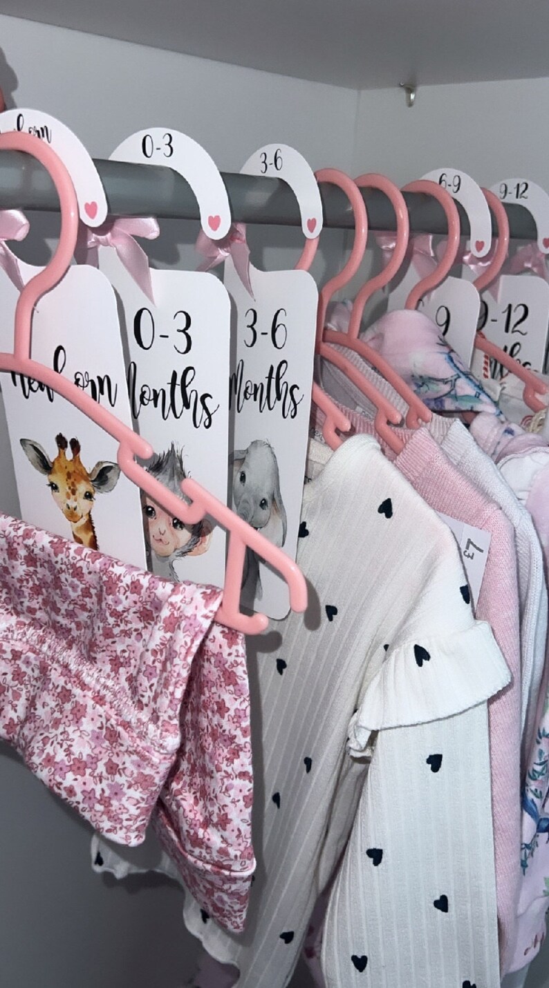 May include: Pink hangers with labels for different age ranges of baby clothes. The labels say "0-3 months", "3-6 months", "6-9 months", and "9-12 months". The hangers are hanging on a metal rod and have baby clothes hanging on them.