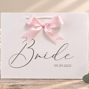 May include: A white gift bag featuring a light pink satin ribbon bow at the top center. The front of the bag displays the word Bride in a dark gray cursive script, with the date 05.09.2025 printed in a smaller font below the text.