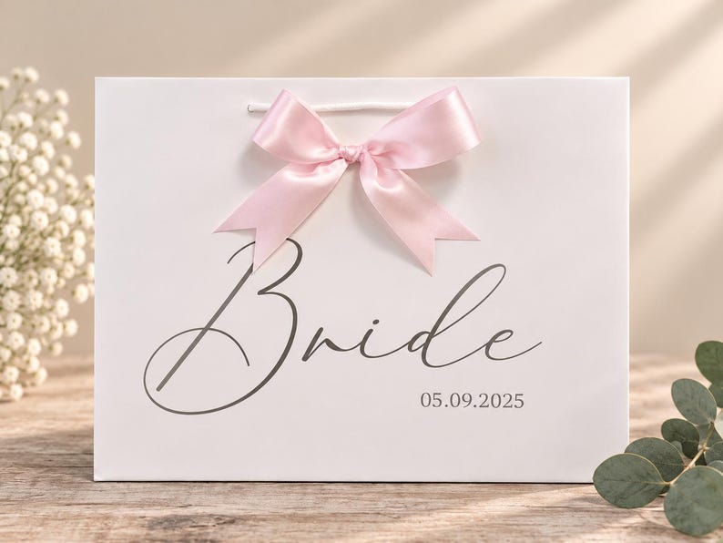 May include: A white gift bag featuring a light pink satin ribbon bow at the top center. The front of the bag displays the word Bride in a dark gray cursive script, with the date 05.09.2025 printed in a smaller font below the text.