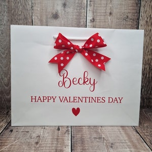 May include: White gift bag with a red polka dot bow and the text "Becky Happy Valentines Day" with a red heart.