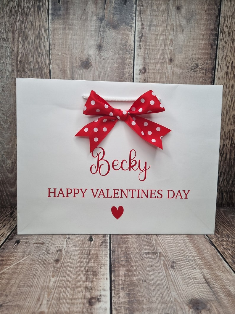 May include: White gift bag with a red polka dot bow and the text "Becky Happy Valentines Day" with a red heart.