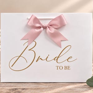 May include: A white rectangular gift bag featuring a dusty pink satin ribbon bow at the top center. The front of the bag displays the text "Bride TO BE" in a metallic gold script and serif font, with white cord handles.