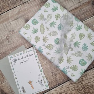 May include: A white gift box with a green and white tropical leaf pattern and a silver ribbon. The box is sitting on a wooden surface. A card with the text "Handmade with love for you!" is next to the box.