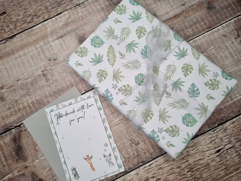 May include: A white gift box with a green and white tropical leaf pattern and a silver ribbon. The box is sitting on a wooden surface. A card with the text "Handmade with love for you!" is next to the box.