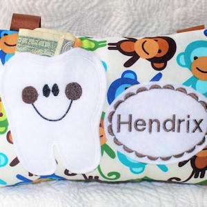 May include: A white tooth-shaped pillow with a smiling face and brown eyes. The pillow has a pocket for a lost tooth and is decorated with a monkey print. The pillow has a personalized embroidered name tag that says "Hendrix".