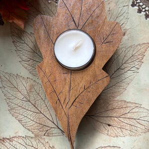 May include: A wooden leaf-shaped tea light holder with a white tea light inside. The holder is carved with leaf veins and sits on a background of pressed leaves.