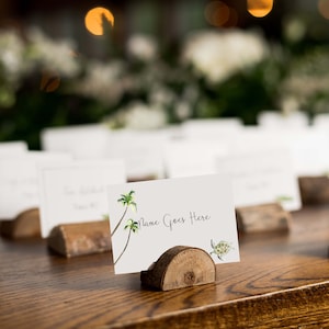 Puede incluir: Tarjetas de lugar blancas con un tema tropical, con palmeras y una tortuga, se muestran en soportes de madera sobre una mesa de madera. Las tarjetas tienen el texto "Name Goes Here" escrito en ellas.
