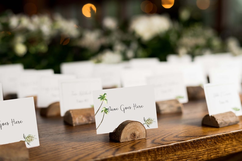 Puede incluir: Tarjetas de lugar blancas con un tema tropical, con palmeras y una tortuga, se muestran en soportes de madera sobre una mesa de madera. Las tarjetas tienen el texto "Name Goes Here" escrito en ellas.