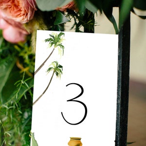 Puede incluir: Tarjeta blanca con el n&uacute;mero "3" en negro y una ilustraci&oacute;n de acuarela de dos palmeras. La tarjeta est&aacute; unida a un clip de metal dorado.