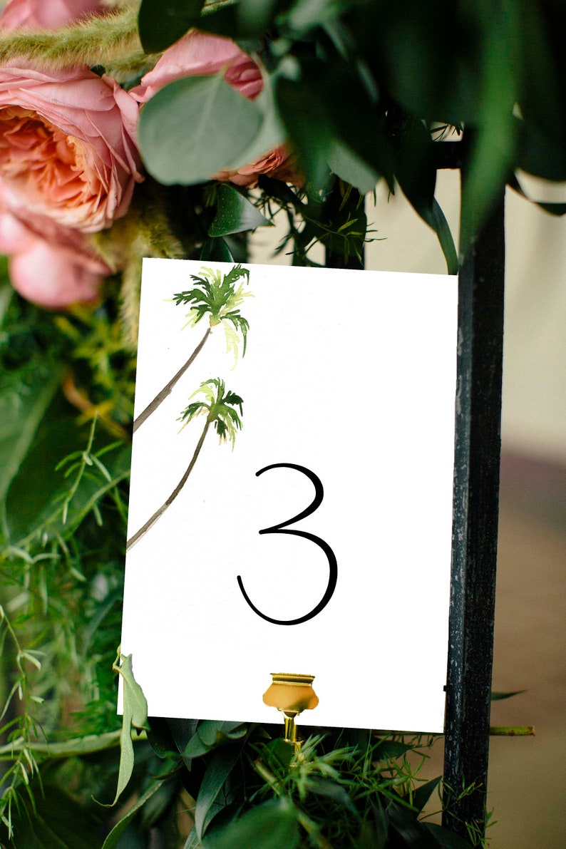 Puede incluir: Tarjeta blanca con el n&uacute;mero "3" en negro y una ilustraci&oacute;n de acuarela de dos palmeras. La tarjeta est&aacute; unida a un clip de metal dorado.