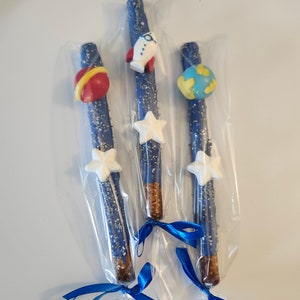 May include: Three blue chocolate-covered pretzel rods with white stars, a red and yellow planet, and a blue and green planet. The pretzels are tied together with blue ribbon.