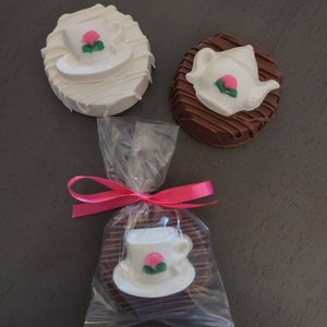 May include: Three chocolate-covered cookies decorated with white chocolate and a teacup, teapot, and flower design. The cookies are packaged in a clear cellophane bag with a pink ribbon.