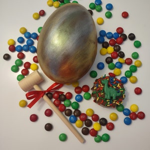 May include: A golden egg with a metallic sheen sits on a white surface surrounded by colorful M&M's candies. A wooden mallet with a red ribbon is next to the egg. A chocolate treat with a green dinosaur and sprinkles is also visible.