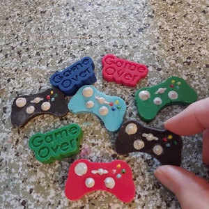 May include: A collection of six colorful game controller shaped soaps with the words "Game Over" on them. The soaps are in various colors including green, blue, red, and brown.