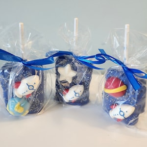 May include: Three blue chocolate-covered marshmallow pops decorated with white fondant rockets, stars, and planets. The pops are wrapped in clear cellophane bags tied with blue ribbon.
