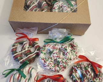May include: A gift box and individual clear bags of chocolate-covered pretzels. The pretzels are decorated with red, green, and white stripes and sprinkles. Each pretzel is tied with a red or green ribbon. The gift box is brown with a clear window.
