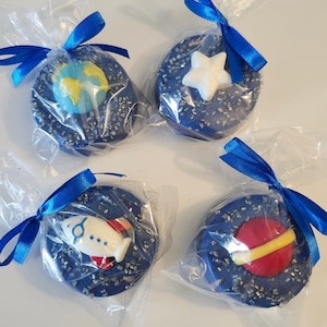 May include: Four individually wrapped chocolate-covered Oreos decorated with space-themed fondant. The Oreos are covered in blue icing and silver sprinkles. The fondant decorations include a rocket, a planet, a star, and a globe.